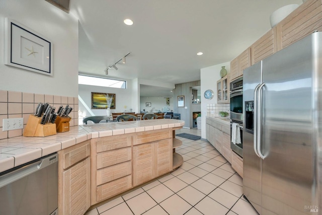 kitchen with tile counters, decorative backsplash, light brown cabinetry, and appliances with stainless steel finishes