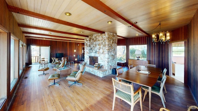 dining space with a notable chandelier, a stone fireplace, light wood-type flooring, beamed ceiling, and wood walls