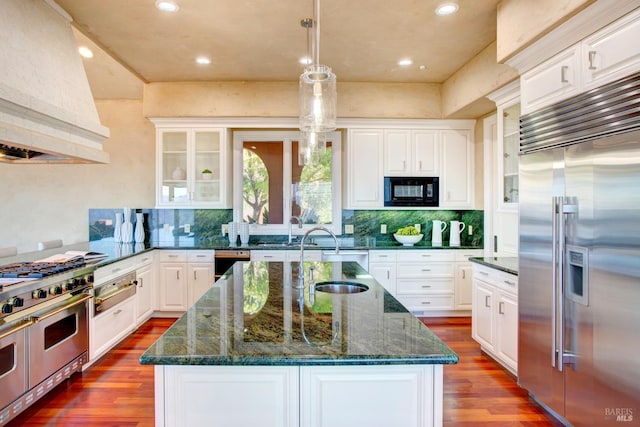 kitchen with premium appliances, an island with sink, glass insert cabinets, premium range hood, and white cabinetry