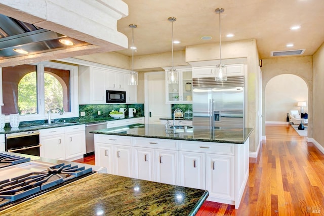 kitchen with arched walkways, white cabinets, an island with sink, and built in appliances