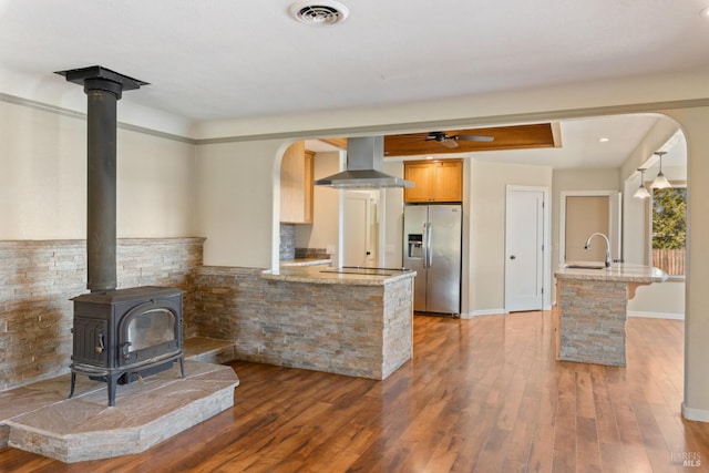 kitchen with a wood stove, wall chimney range hood, hardwood / wood-style flooring, stainless steel fridge, and kitchen peninsula