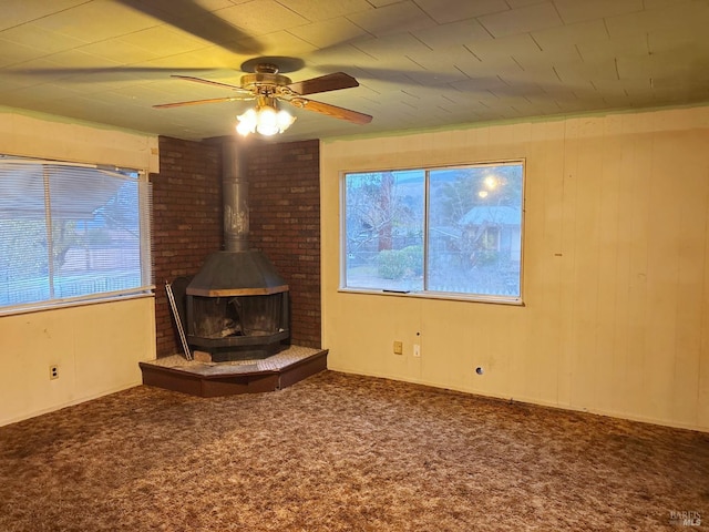 unfurnished living room featuring a wood stove, ceiling fan, wooden walls, and carpet floors