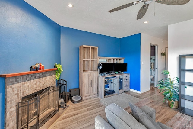 living room with a tiled fireplace, ceiling fan, a textured ceiling, and light wood-type flooring