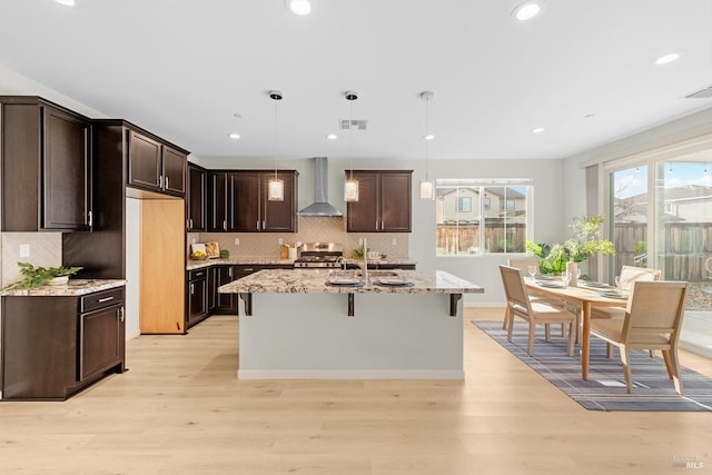 kitchen with decorative light fixtures, wall chimney range hood, a center island with sink, light hardwood / wood-style flooring, and light stone countertops