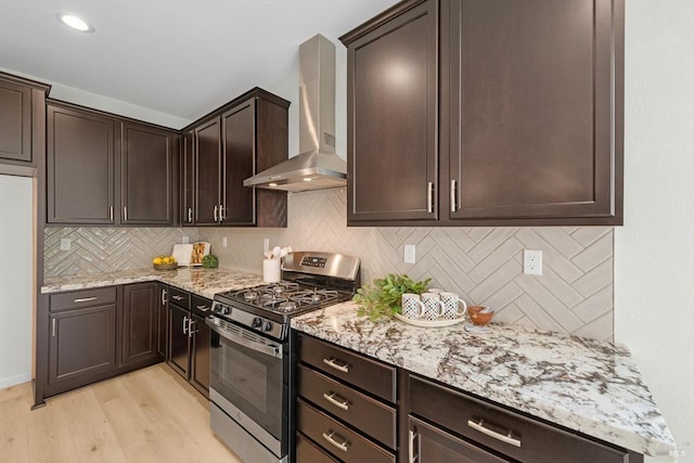 kitchen with stainless steel gas range oven, tasteful backsplash, dark brown cabinetry, light stone counters, and wall chimney exhaust hood