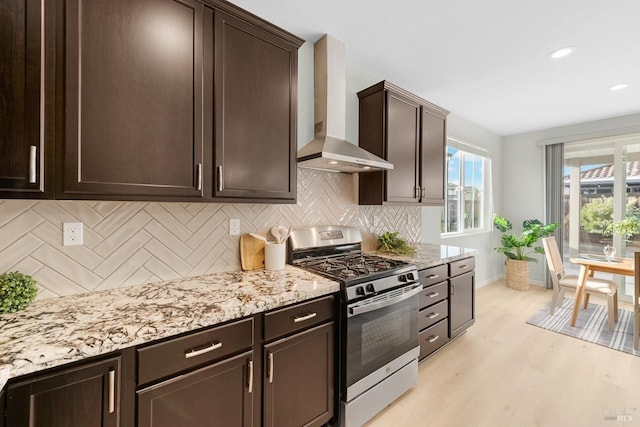 kitchen with stainless steel gas range oven, light wood-style floors, dark brown cabinetry, wall chimney exhaust hood, and a healthy amount of sunlight