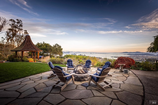 patio terrace at dusk featuring a gazebo, a fire pit, a water view, and a lawn