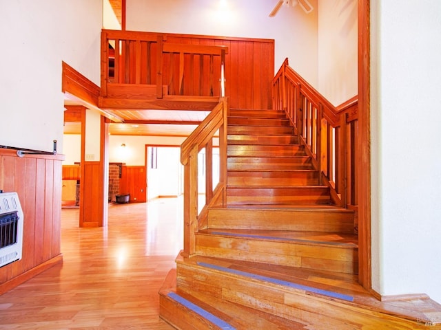 staircase featuring a high ceiling, heating unit, ceiling fan, hardwood / wood-style floors, and wood walls