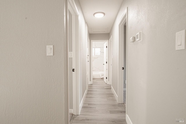 hallway featuring attic access, baseboards, a textured wall, and light wood finished floors