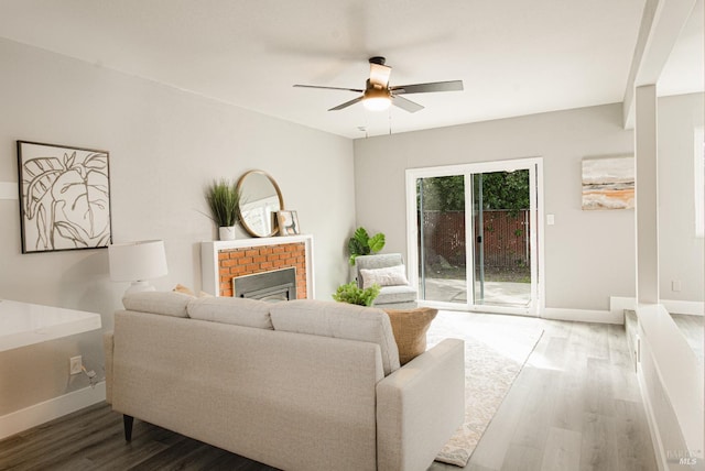 living area featuring a brick fireplace, ceiling fan, baseboards, and wood finished floors