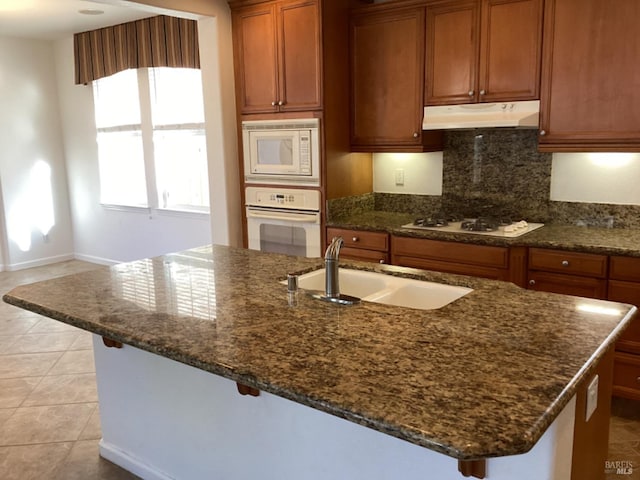 kitchen with a kitchen island with sink, sink, white appliances, tasteful backsplash, and dark stone countertops