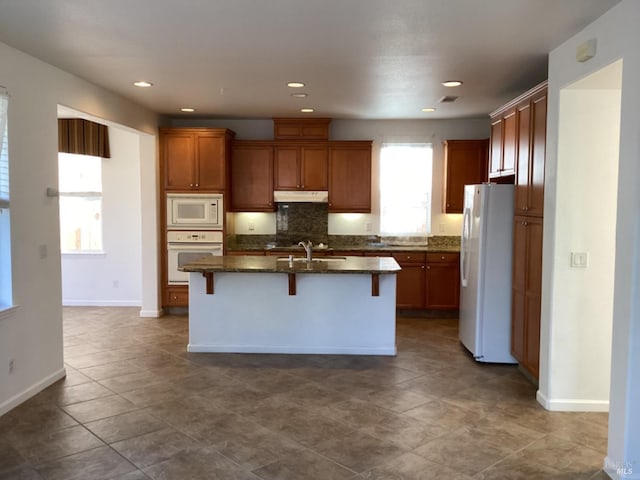 kitchen with white appliances, a center island with sink, dark stone counters, and a breakfast bar area