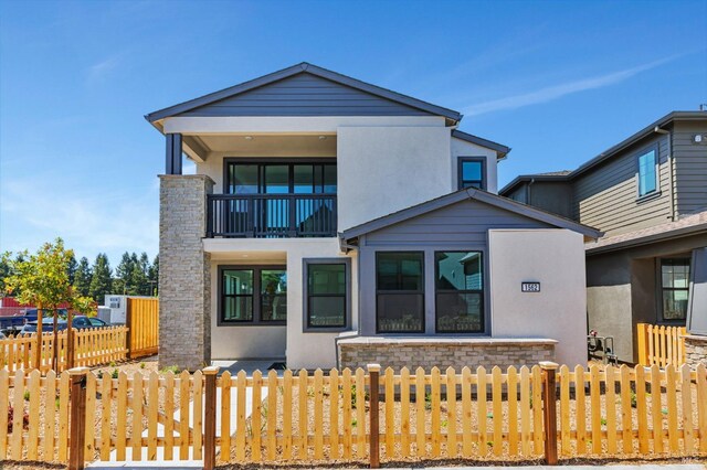 back of house at dusk with fence, a balcony, stone siding, and stucco siding