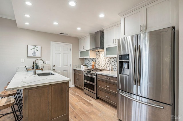 kitchen with light stone countertops, wall chimney range hood, light hardwood / wood-style floors, appliances with stainless steel finishes, and sink