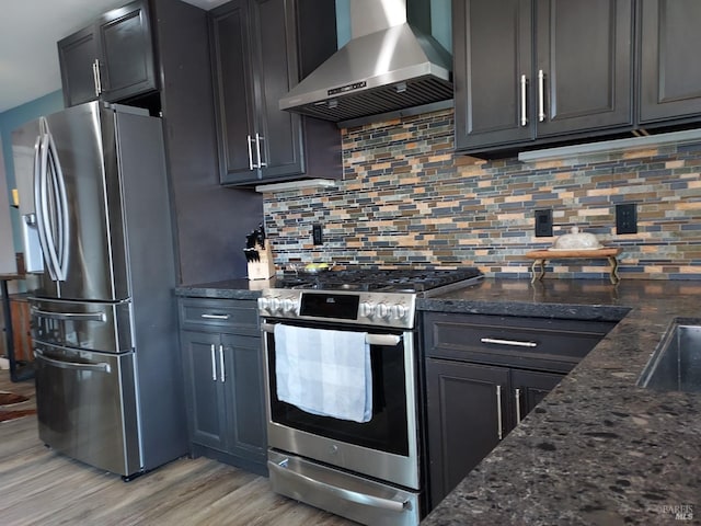 kitchen featuring wall chimney exhaust hood, dark stone countertops, light hardwood / wood-style floors, decorative backsplash, and appliances with stainless steel finishes