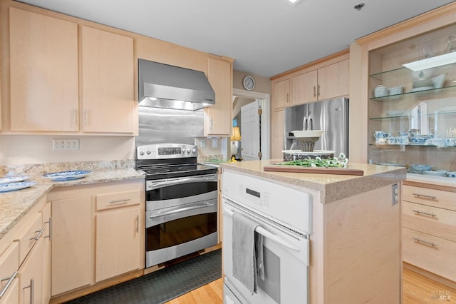 kitchen with light brown cabinets, wall chimney range hood, light stone counters, light hardwood / wood-style flooring, and appliances with stainless steel finishes