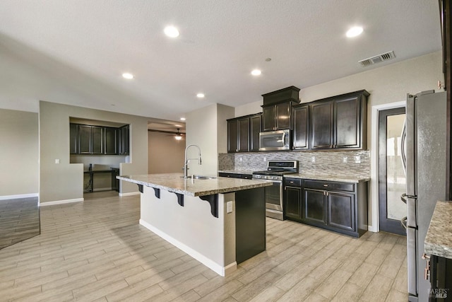 kitchen with sink, light hardwood / wood-style flooring, an island with sink, appliances with stainless steel finishes, and light stone counters