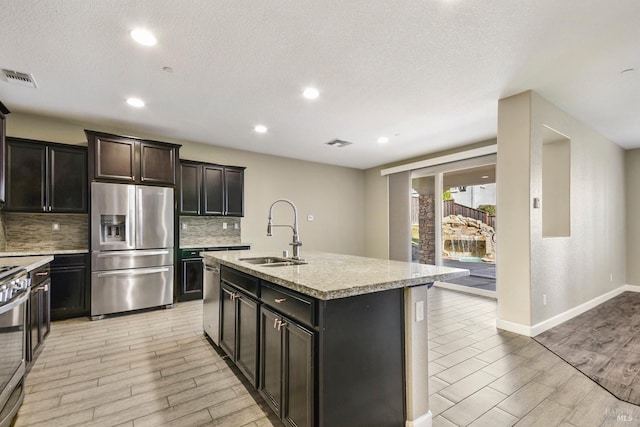 kitchen with light stone countertops, appliances with stainless steel finishes, backsplash, a kitchen island with sink, and sink