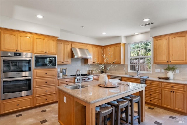 kitchen featuring light stone counters, built in microwave, double oven, a center island with sink, and range hood