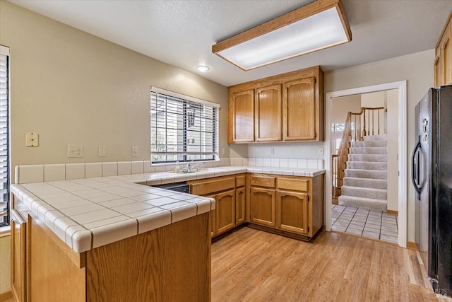 kitchen with tile countertops, dishwasher, black fridge, light hardwood / wood-style floors, and kitchen peninsula