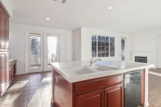 kitchen featuring a kitchen island with sink, sink, light tile patterned floors, black dishwasher, and tile counters