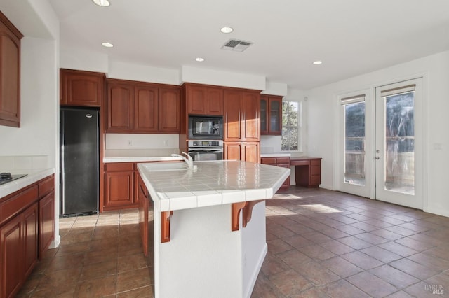 kitchen featuring stainless steel oven, a kitchen island with sink, refrigerator, tile counters, and black microwave