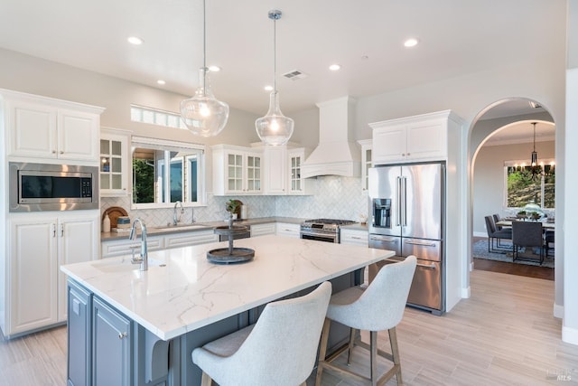 kitchen featuring light stone countertops, white cabinetry, an island with sink, appliances with stainless steel finishes, and custom exhaust hood