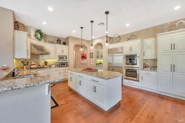 kitchen featuring pendant lighting, built in appliances, decorative backsplash, sink, and range hood