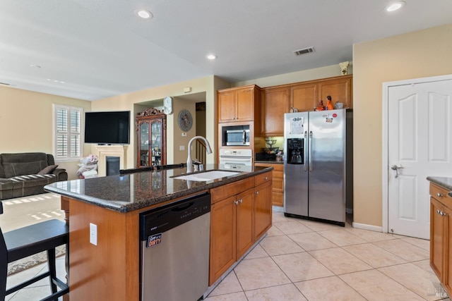 kitchen with sink, light tile patterned floors, dark stone countertops, appliances with stainless steel finishes, and an island with sink