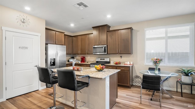 kitchen with a kitchen bar, light stone counters, stainless steel appliances, light hardwood / wood-style flooring, and an island with sink