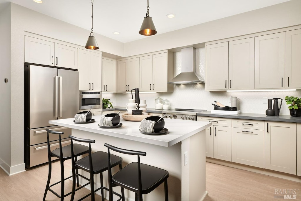 kitchen with appliances with stainless steel finishes, wall chimney exhaust hood, pendant lighting, light hardwood / wood-style flooring, and a kitchen island