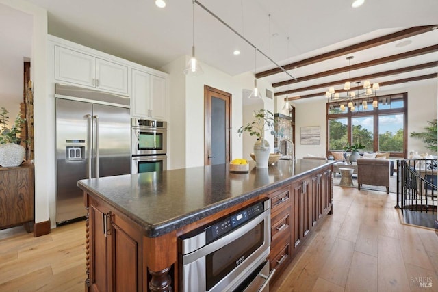 kitchen with brown cabinets, a center island with sink, dark countertops, light wood-style flooring, and appliances with stainless steel finishes