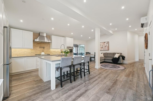 kitchen with white cabinets, appliances with stainless steel finishes, wall chimney exhaust hood, light wood-type flooring, and a center island with sink