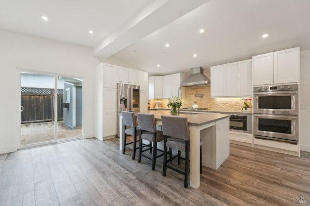 kitchen featuring wall chimney exhaust hood, white cabinetry, stainless steel appliances, an island with sink, and a breakfast bar area