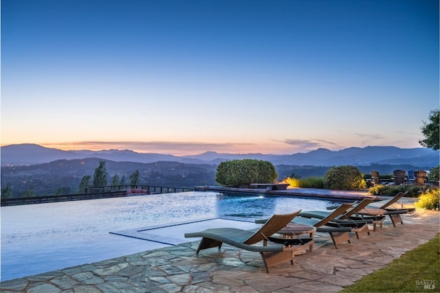 pool at dusk featuring an infinity pool, a patio, fence, and a mountain view