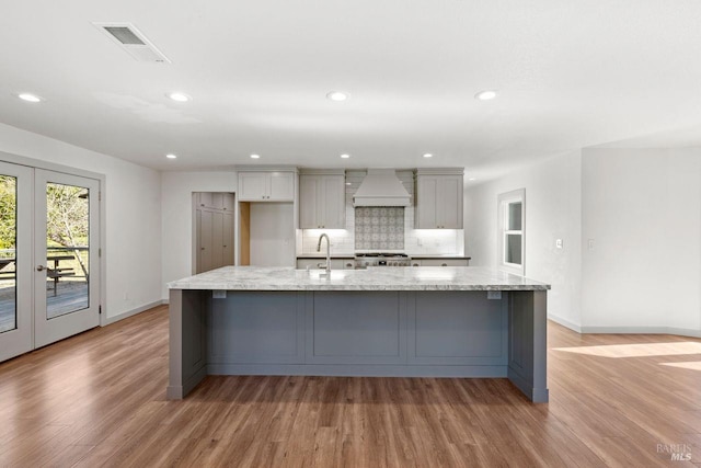 kitchen with light stone counters, light wood-type flooring, a spacious island, custom exhaust hood, and decorative backsplash