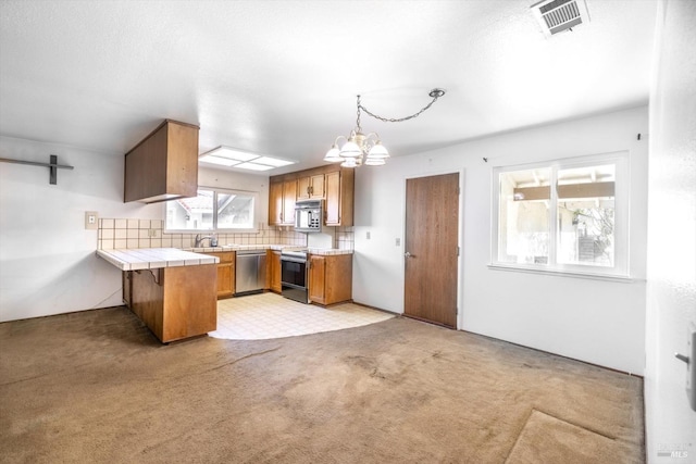 kitchen with tile counters, brown cabinetry, appliances with stainless steel finishes, decorative light fixtures, and a peninsula