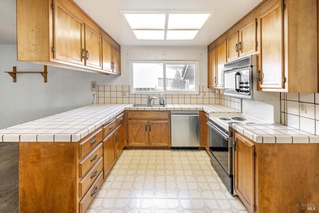 kitchen featuring brown cabinets, tile countertops, light floors, backsplash, and appliances with stainless steel finishes