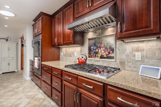 kitchen with tasteful backsplash, wall chimney range hood, stainless steel appliances, light tile patterned floors, and light stone counters