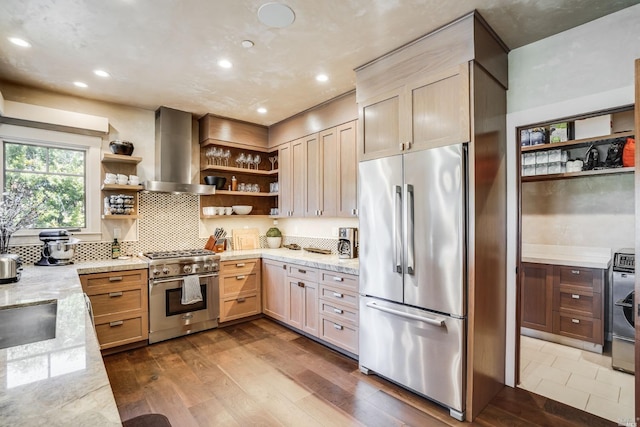 kitchen featuring appliances with stainless steel finishes, light brown cabinetry, wood-type flooring, wall chimney range hood, and light stone counters