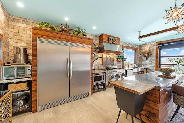 kitchen with wall chimney exhaust hood, butcher block countertops, light wood-style flooring, built in appliances, and vaulted ceiling