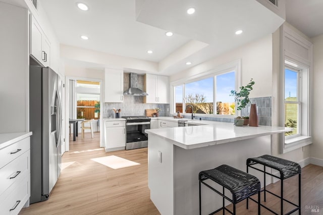 kitchen featuring stainless steel appliances, a peninsula, white cabinets, wall chimney exhaust hood, and a kitchen bar