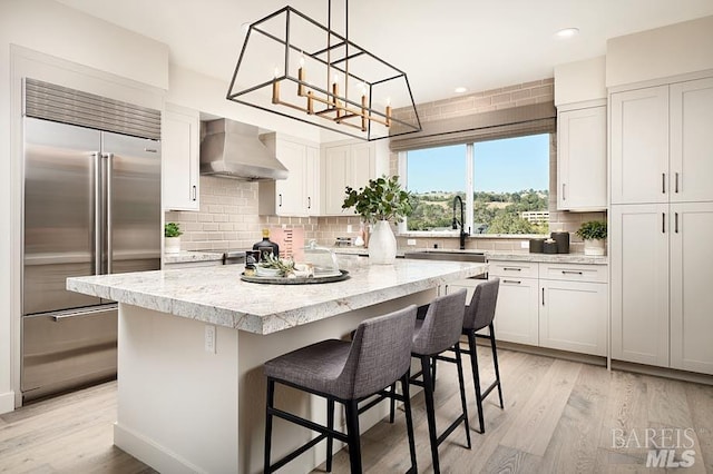 kitchen featuring a kitchen island, white cabinets, stainless steel built in refrigerator, and wall chimney range hood