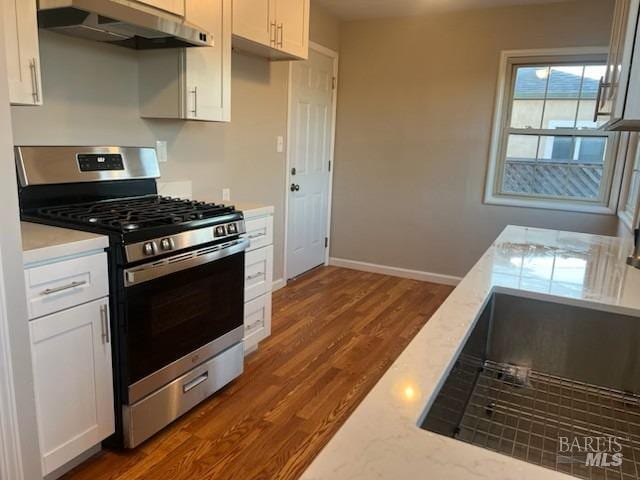 kitchen with light stone counters, stainless steel gas range oven, under cabinet range hood, white cabinetry, and dark wood-type flooring