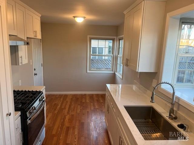 kitchen with dark wood finished floors, under cabinet range hood, white cabinets, a sink, and stainless steel range with gas cooktop