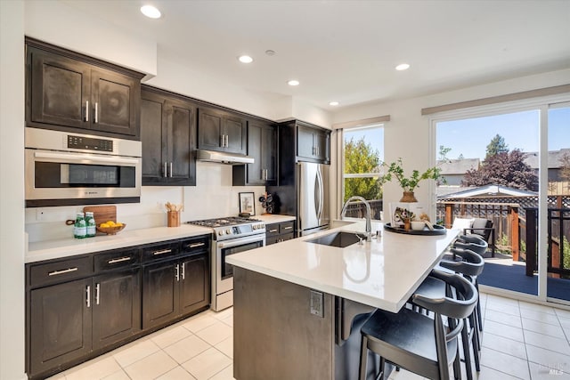 kitchen with an island with sink, a breakfast bar area, stainless steel appliances, dark brown cabinetry, and sink