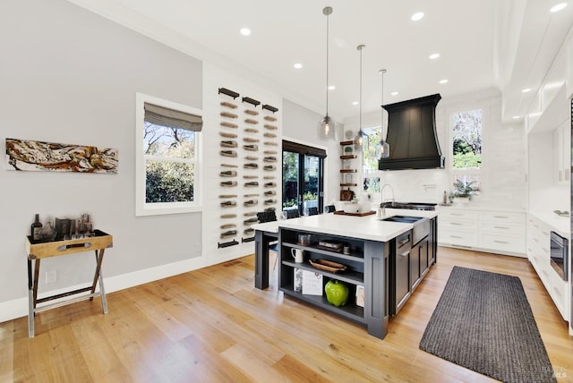 kitchen with custom exhaust hood, an island with sink, light wood-type flooring, hanging light fixtures, and white cabinets