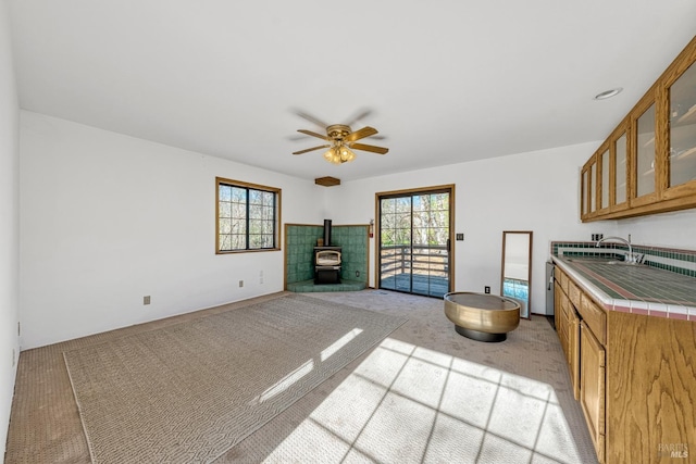 kitchen featuring light carpet, ceiling fan, tile counters, a wood stove, and sink