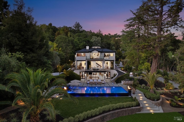back of house at dusk featuring stairway, an outdoor pool, a balcony, a lawn, and a patio