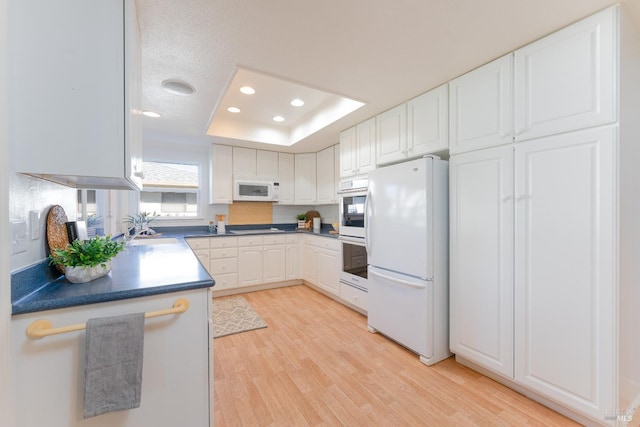 kitchen with white appliances, a raised ceiling, light wood-style flooring, white cabinetry, and recessed lighting